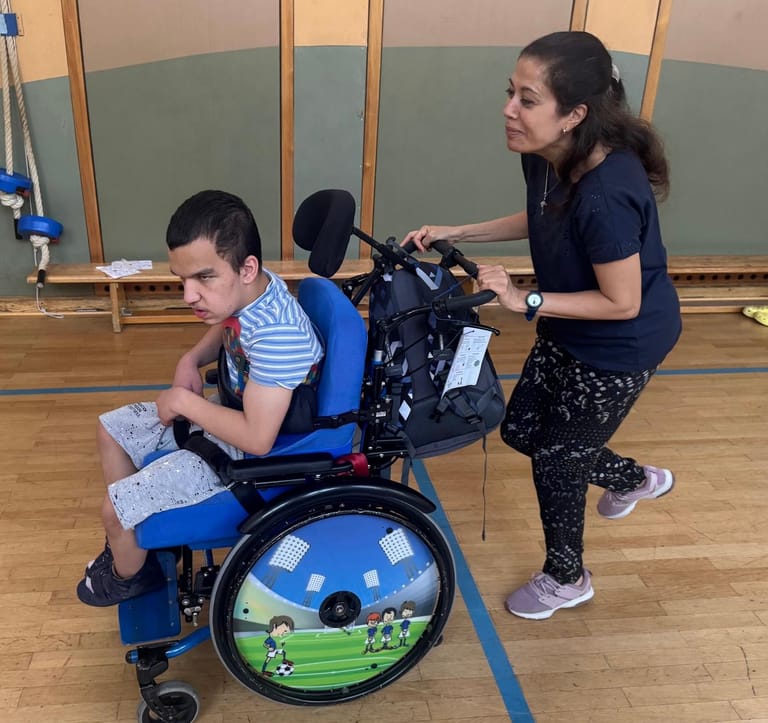 Frau schiebt Kind im Rollstuhl durch den Turnsaal.