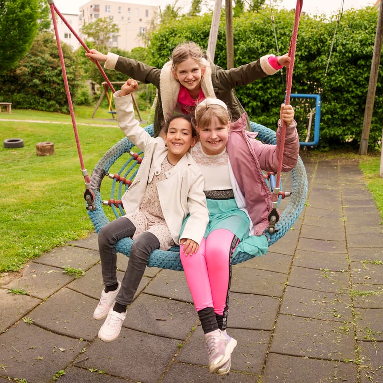 Drei Kinder schaukeln auf der Korbschaukel und lächeln.